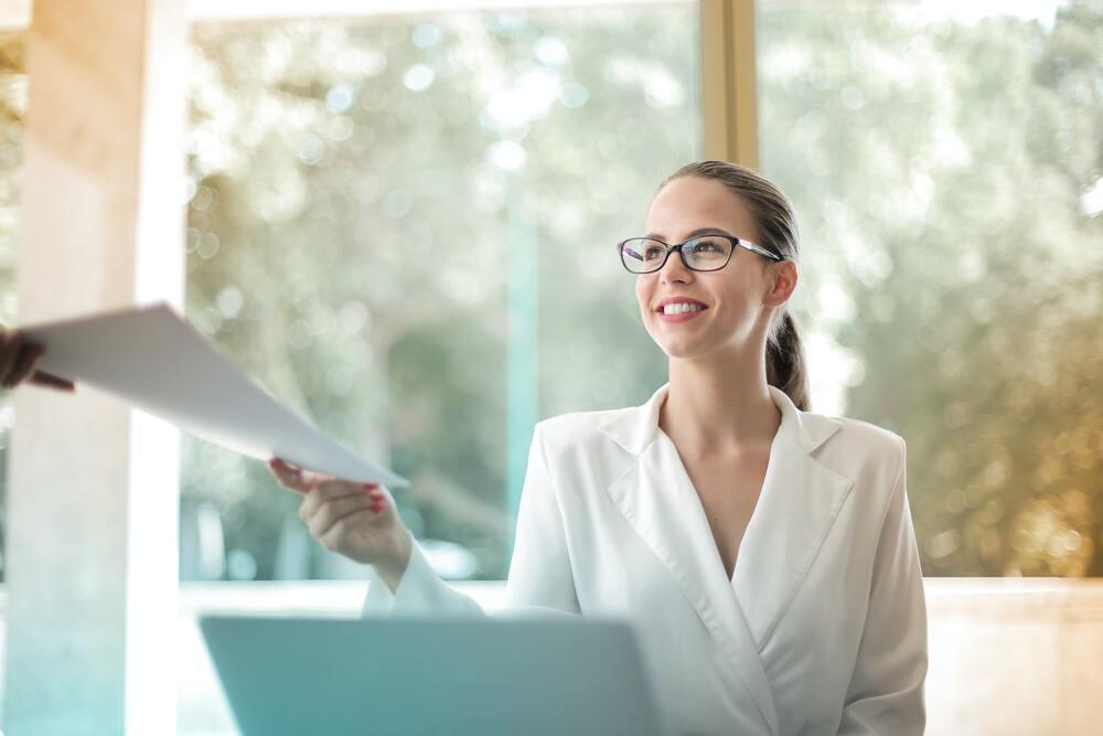 Free Low angle of successful female executive manager in classy style sitting at table with laptop in contemporary workplace and passing documents to colleague